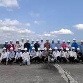 A large group of people in hard hats pose for a photo outdoors, with a body of water and hills in the background.