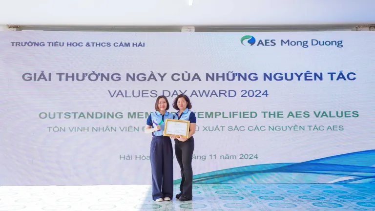 Two women stand on a stage in front of a large screen displaying the text "Values Day Award 2024." Both are dressed in matching blue uniforms with light blue scarves and hold a framed certificate and microphone. The screen also shows AES' Mong Duong logo and Vietnamese text highlighting the recognition event.