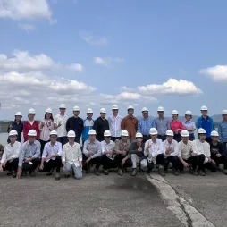A large group of people in hard hats pose for a photo outdoors, with a body of water and hills in the background.